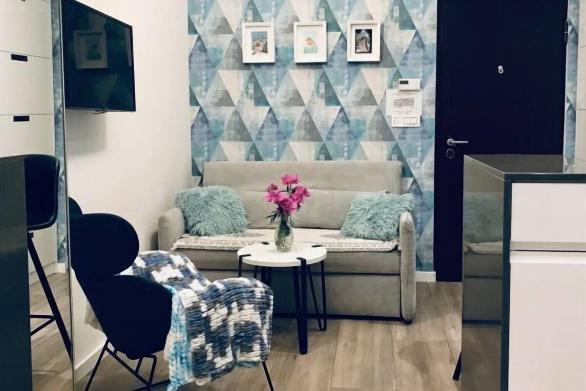 A modern living room with a sofa, a small table with a vase of flowers, a black chair, and a TV mounted on the wall.