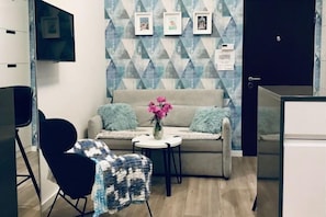 A modern living room with a sofa, a small table with a vase of flowers, a black chair, and a TV mounted on the wall.
