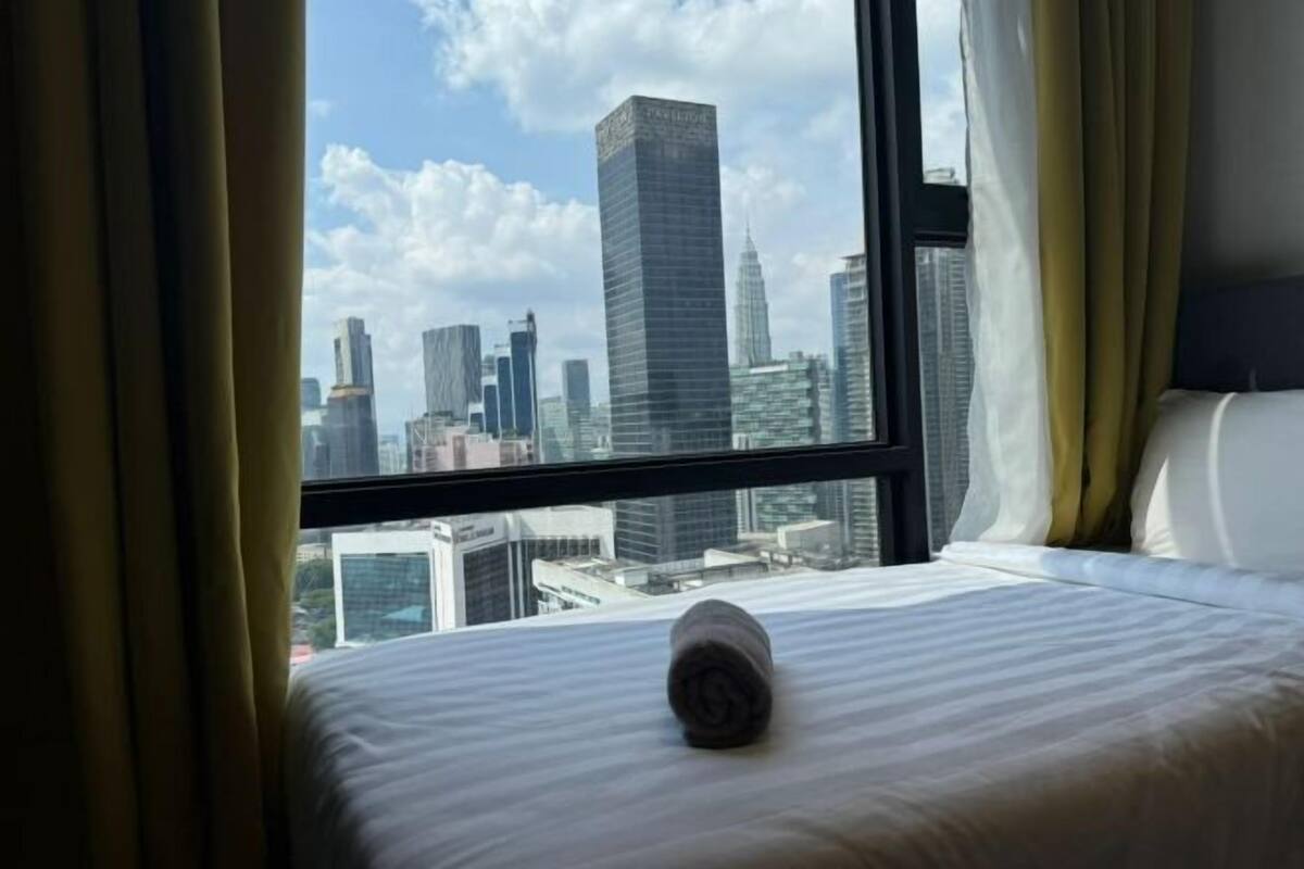 A hotel room with a bed, a rolled towel, and a view of a cityscape through a large window.