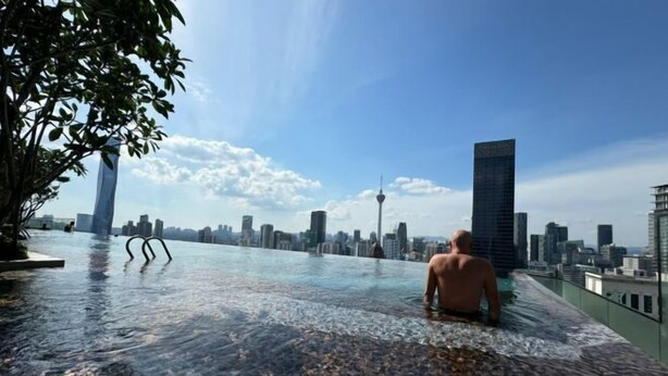 Un hombre sentado en una piscina infinita en la azotea, con el horizonte de la ciudad al fondo.