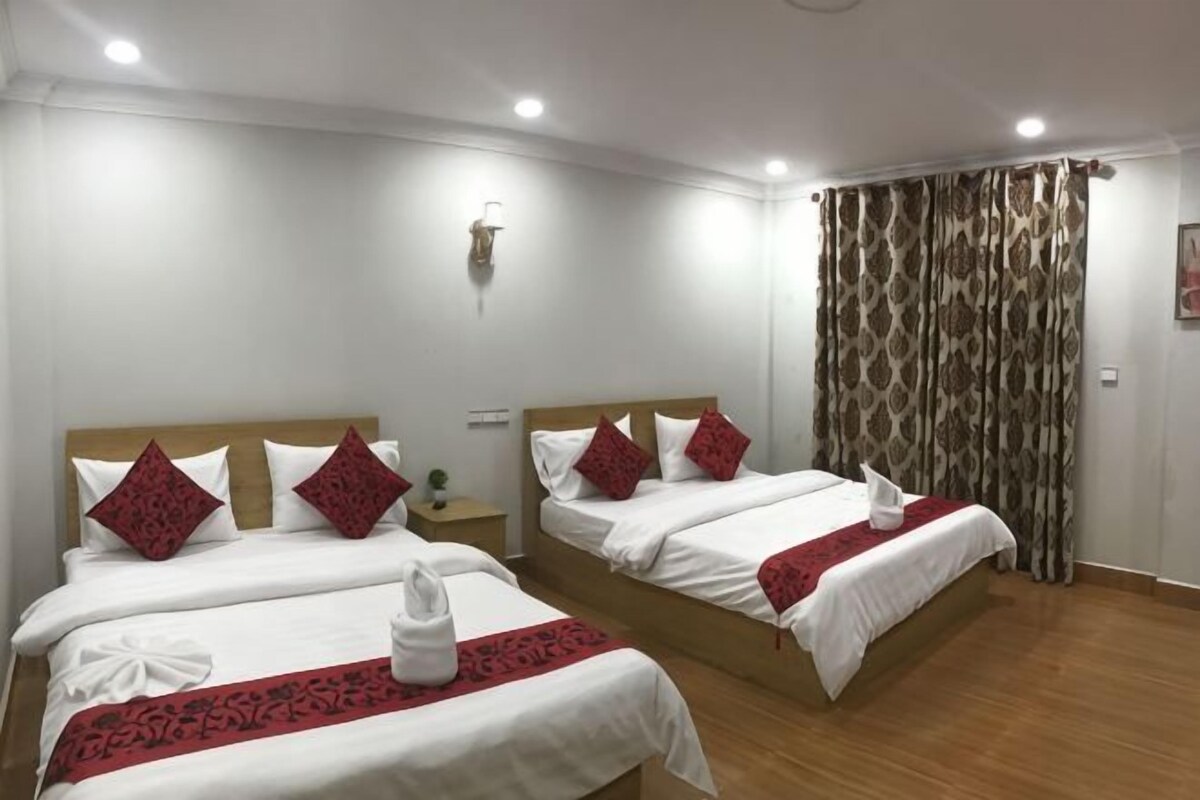 A hotel room with two beds, each with white bedding and red pillows, a wooden headboard, a bedside table with a lamp, and a curtain with a patterned design.