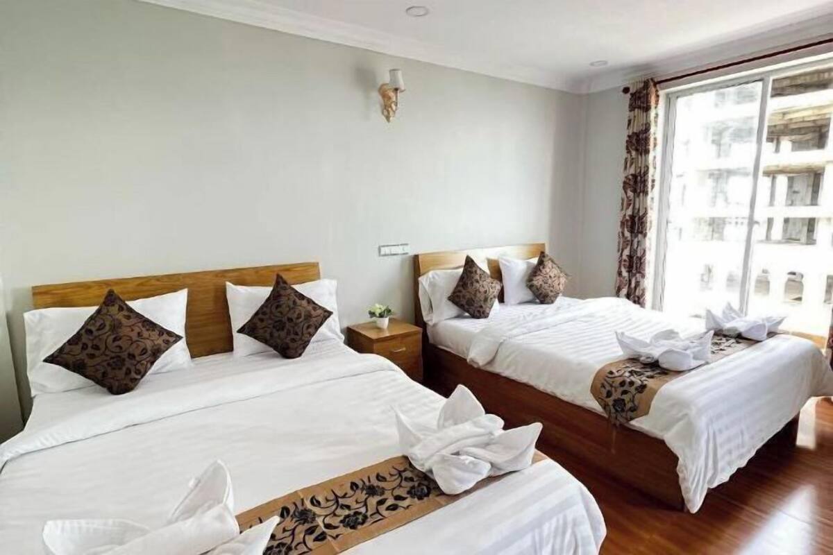 A hotel room with two beds, a nightstand, a wall-mounted lamp, and a large window with curtains.
