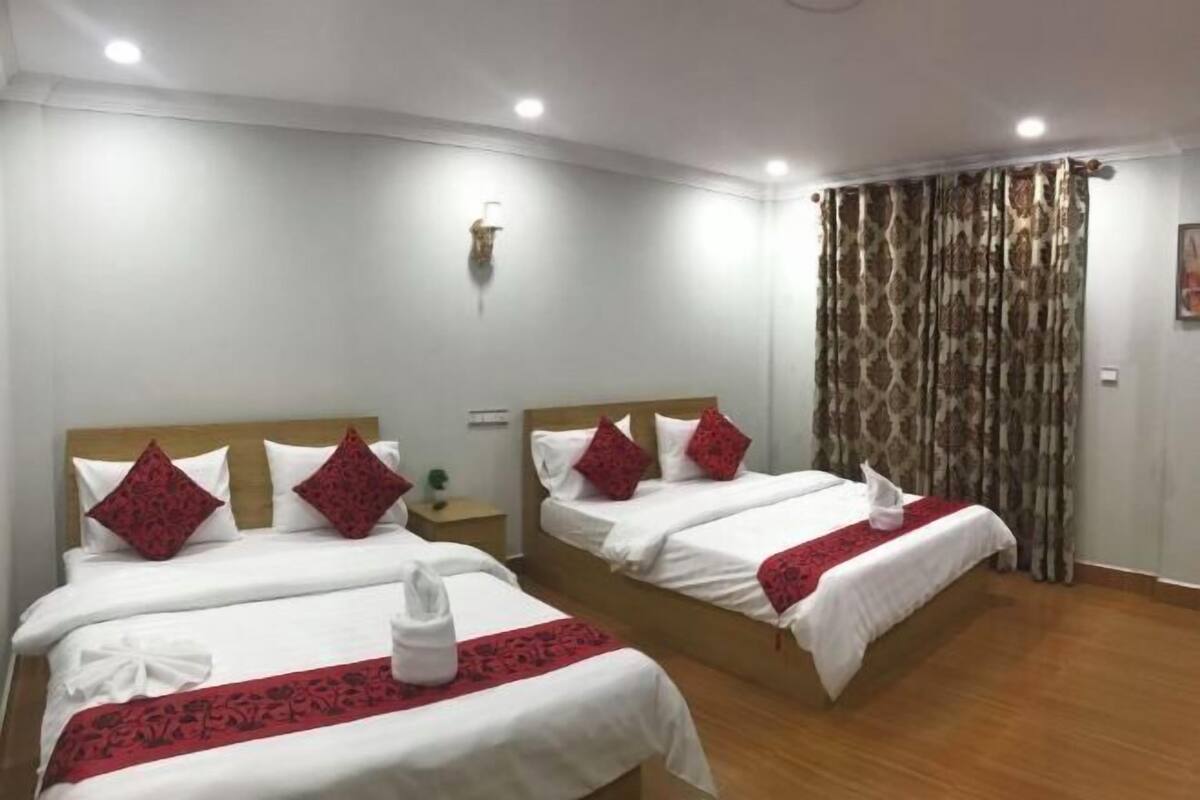 A hotel room with two beds, each with white bedding and red accents, a wooden headboard, a nightstand, and a curtain with a patterned design.
