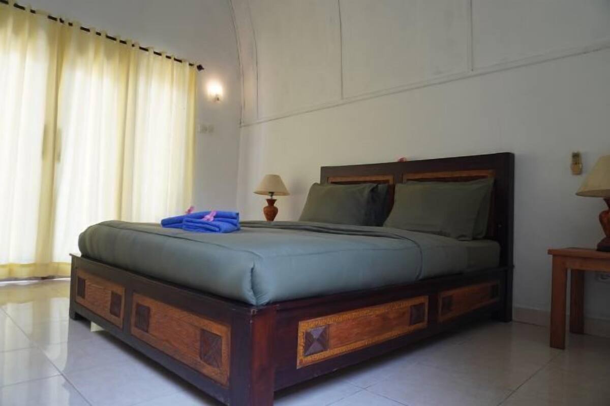 Uma cama com estrutura de madeira e colchão verde, uma mesa de cabeceira com abajur e uma janela com cortinas.