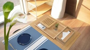 Une chambre d’hôtel avec un set de table à rayures bleues, un vase blanc contenant une seule fleur, un plateau en bois avec une boîte de mouchoirs et une poubelle blanche.