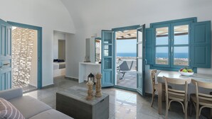 A modern living room with a view of the sea, a dining area with a table and chairs, and a cozy seating area with a sofa and a decorative table. - GTK Santorini Oia Sunset Villas (Santorini)