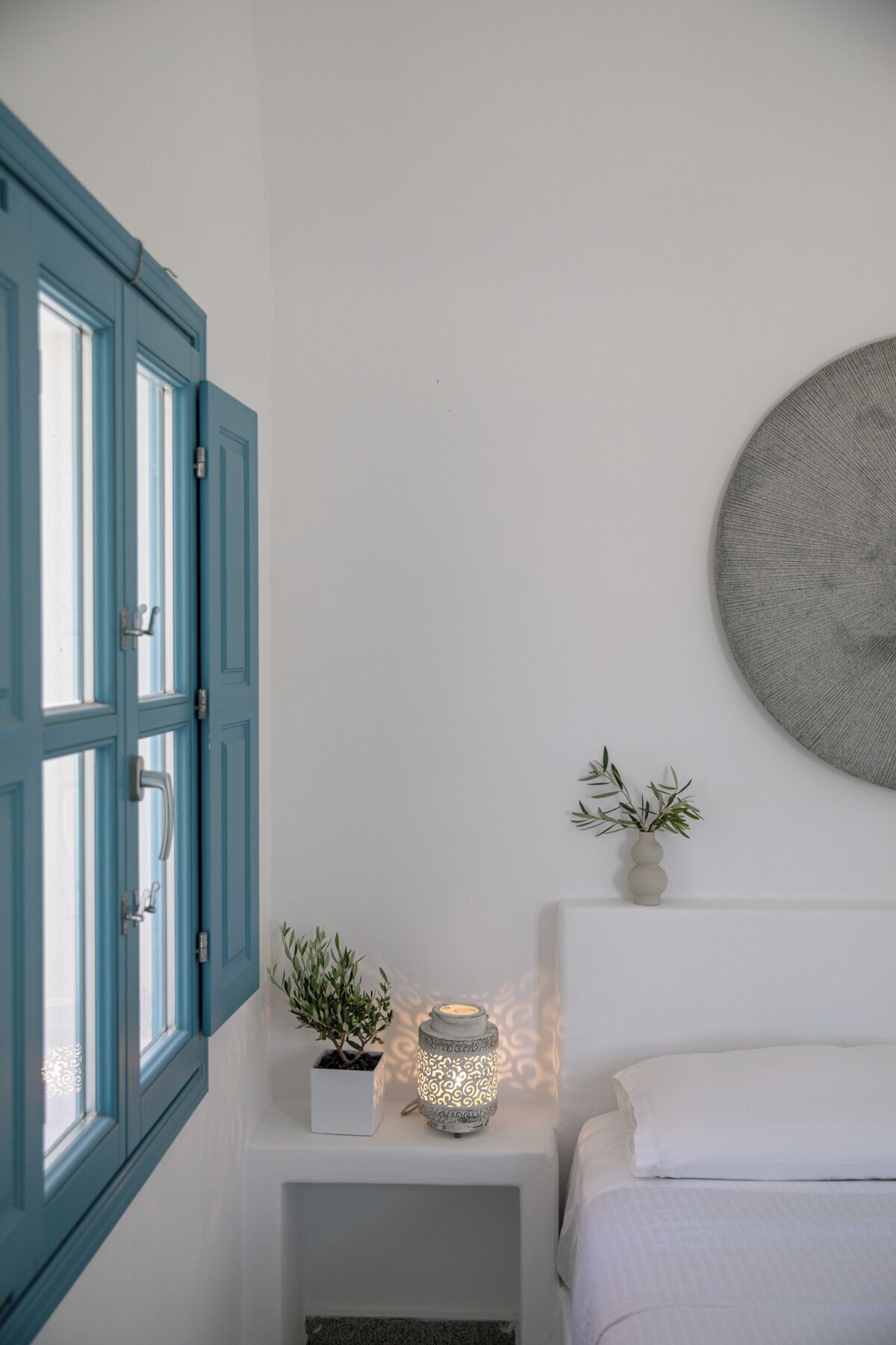 A bedroom with a white bed, a blue window, a small bedside table with a plant and a lantern, and a round wall decoration.