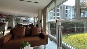 A modern living room with a brown leather sofa, large windows, and a view of a residential building. - Peaceful Stay @MT Khlong Luang (Khlong Luang)