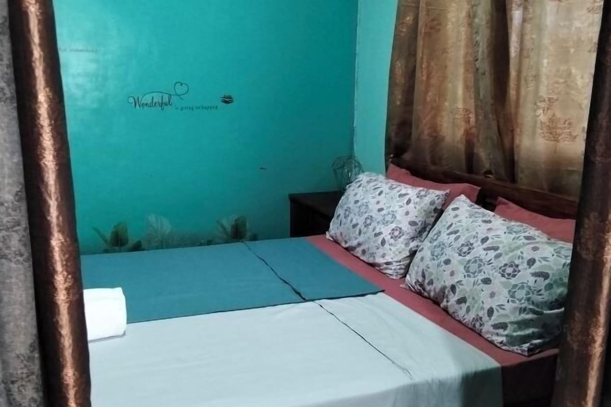 Un letto con cuscini floreali, una parete color petrolio con un cartello che riporta la scritta 'Wonderful' e una tenda.