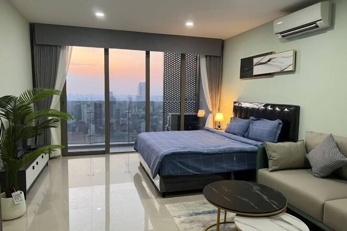 A modern bedroom with a large bed, a sofa, a small table, and a city view through large windows.