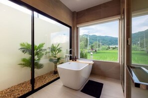 A modern bathroom with a freestanding bathtub, a glass shower enclosure, and a view of a green landscape. - Vann House Chiang Rai- Near Mae Fah Luang University (Chiang Rai)