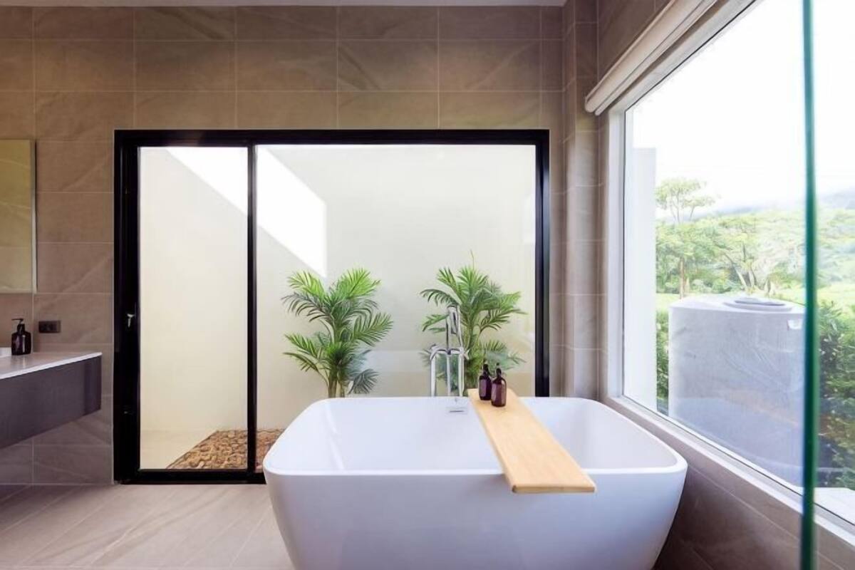 A modern bathroom with a large bathtub, a glass shower enclosure, and a view of greenery outside.