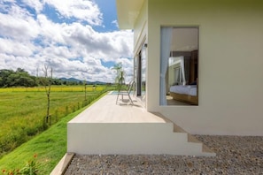 A modern building with a balcony overlooking a green field, a clear sky, and a visible bedroom with a bed and window.