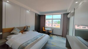 A hotel room with a bed, a small table, a chair, a TV, and a window with a city view. - Ming Hua International Hotel (Boten)