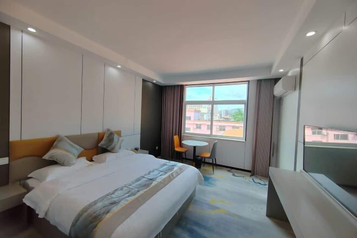 A hotel room with a bed, a small table, a chair, a TV, and a window with a city view.