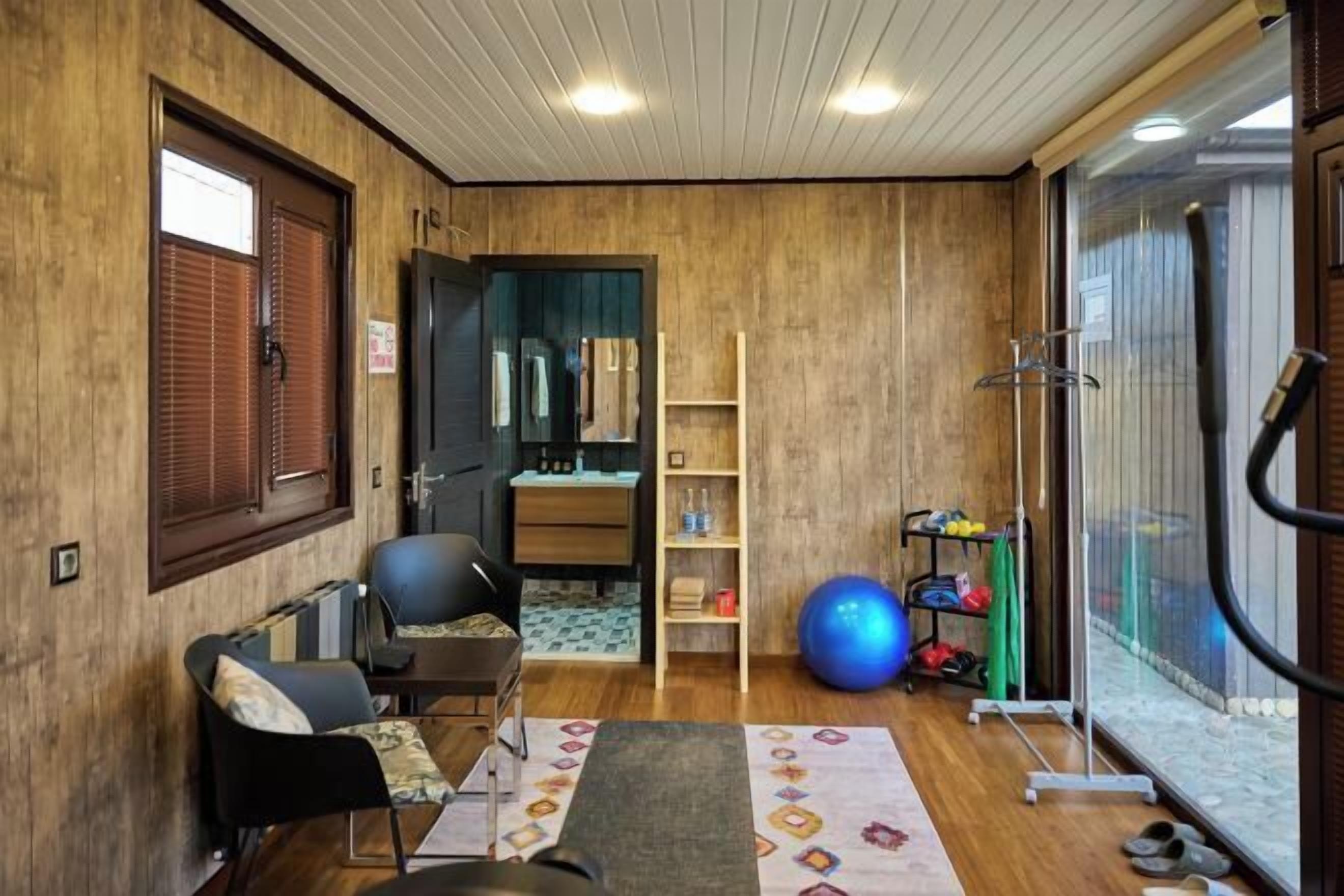 A room with a wooden floor, a black chair, a desk, a blue exercise ball, a shoe rack, and a window with blinds.