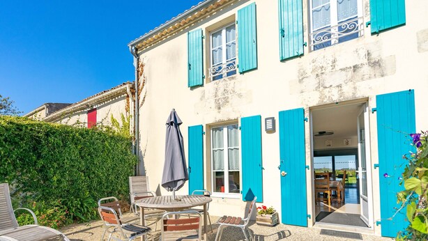 A building with a patio area featuring a table and chairs, a door, and a window with blue shutters.