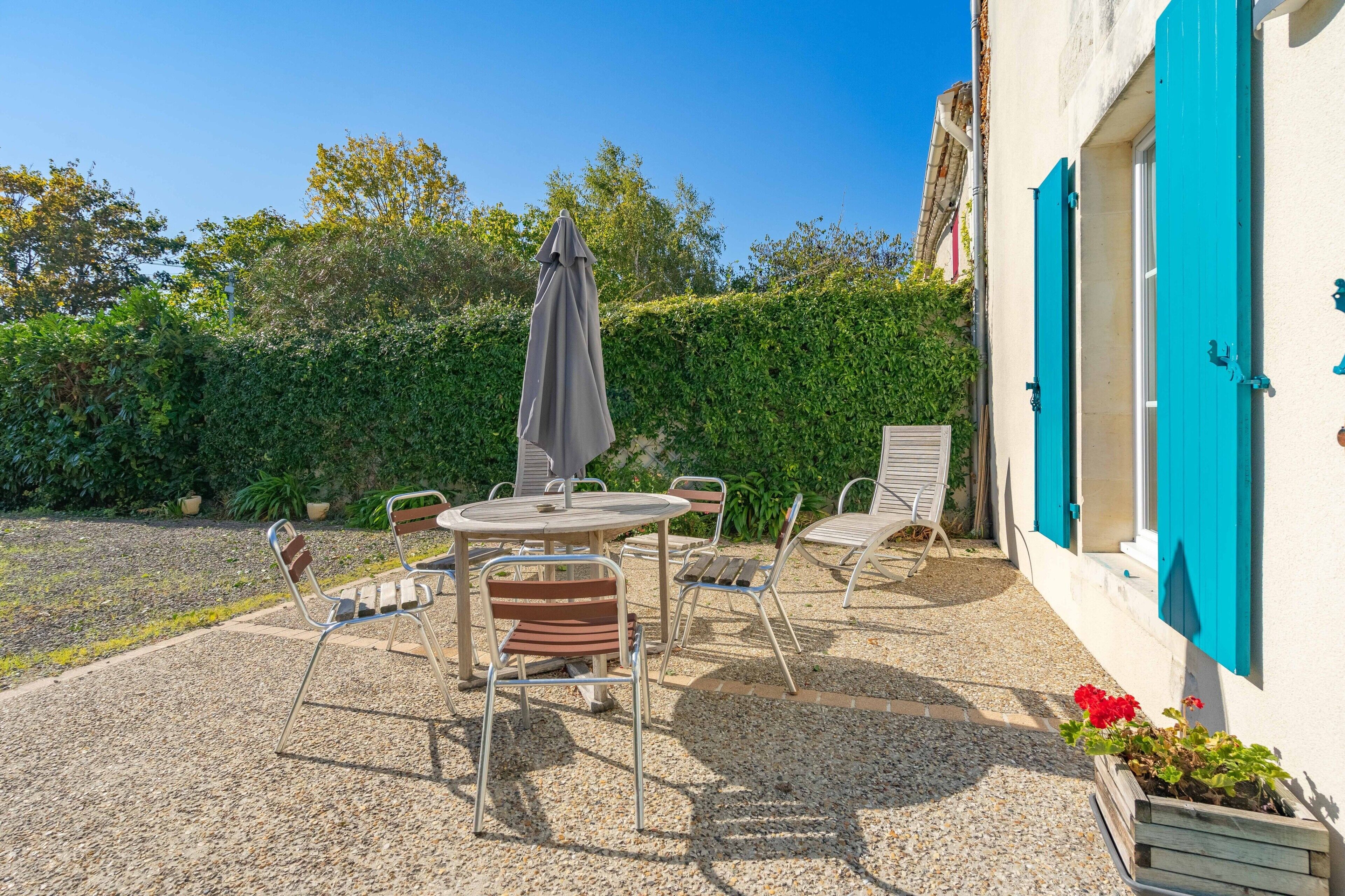 Een terras met een ronde tafel en stoelen, een grijze parasol en een wit gebouw met blauwe luiken.