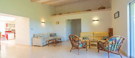 A spacious living room with a high ceiling, exposed beams, a brick wall, and a kitchen in the background.