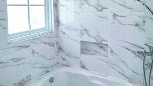 A bathroom with a bathtub, a window, and marble walls. - TwoSpaces Living at Arfina Residence Syariah, Lamongan (Paciran)