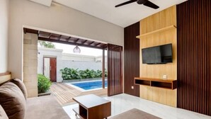 A modern living room with a sofa, coffee table, and a TV mounted on a wooden wall. There is a sliding door leading to a pool area. - The Umah Pandawa 2 (Bali)