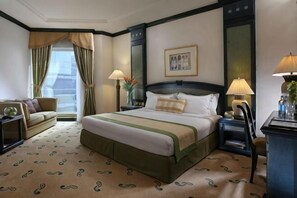 Ein Hotelzimmer mit einem großen Bett, einem Schreibtisch, einem Stuhl, einer Lampe, einem Nachttisch, einem Fenster mit Vorhängen und einem gerahmten Bild an der Wand.