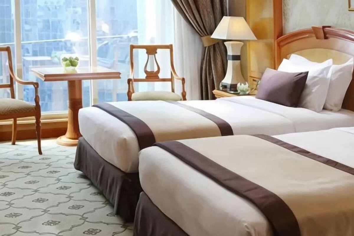 A hotel room with two beds, a small table with a plant, a chair, and a window with a city view.