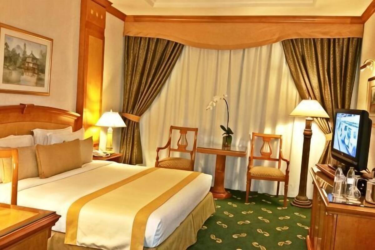 A hotel room with a bed, two lamps, a television, a chair, a small table, and a decorative plant.