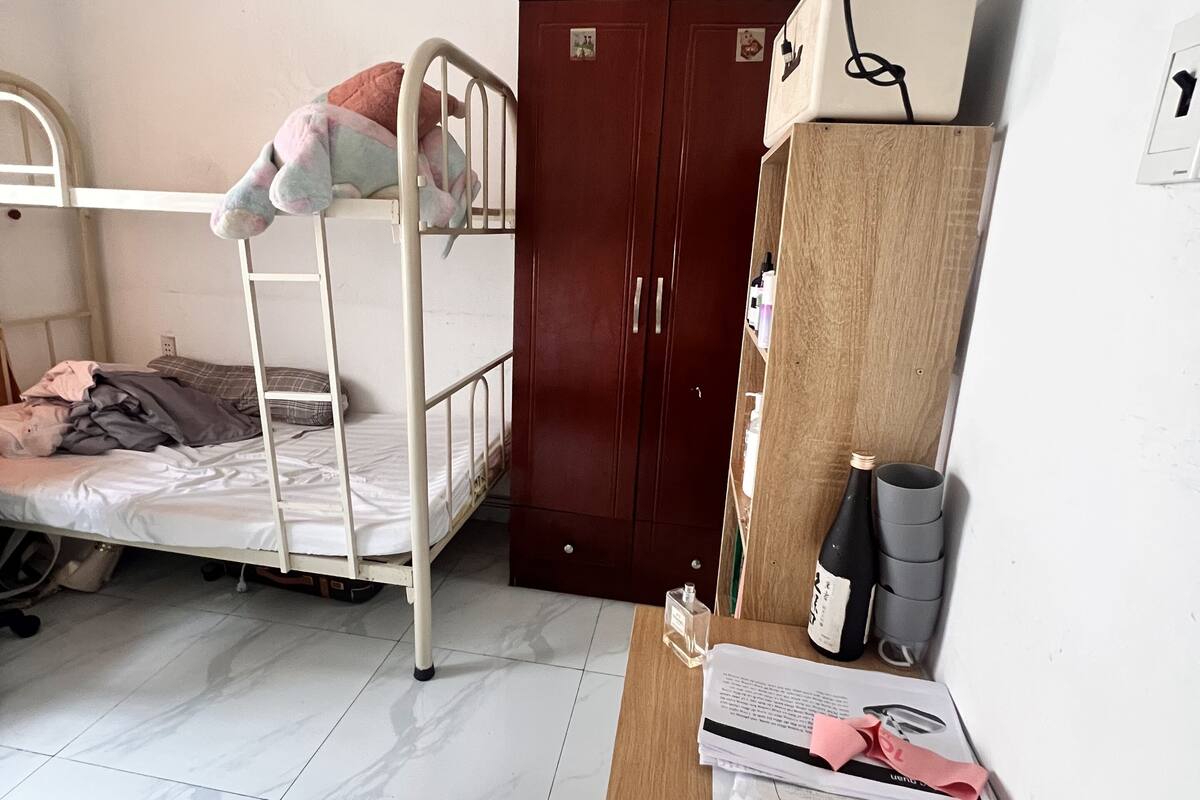 A dormitory room with a bunk bed, a wooden desk with papers and a bottle, a wardrobe, and a wall-mounted coat rack.