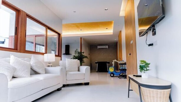 A modern living room with a white sofa, a small armchair, a television mounted on the wall, and a toy car. - Holmestay Hatyai (Hat Yai)