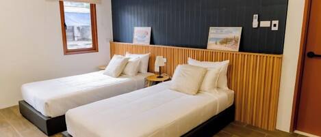 Habitación de hotel con dos camas, cabecera de madera, mesita de noche pequeña con lámpara y una ventana.