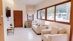 A modern living room with two white sofas, a small side table, a TV mounted on the wall, and large windows offering a view of the outside. - Holmestay Hatyai (Hat Yai)