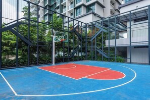 Uma quadra de basquete interna com piso azul e vermelho, cercada por uma grade de metal e painéis de vidro, com edifícios ao fundo.