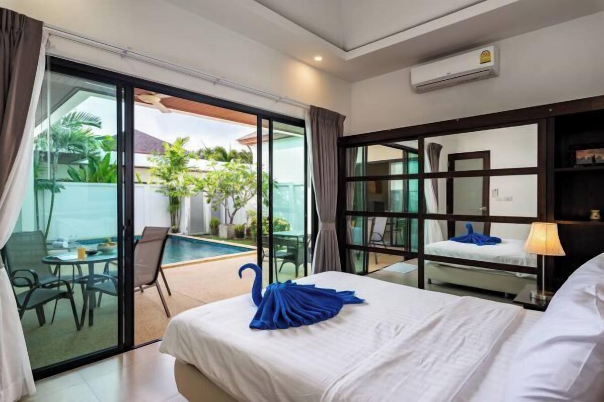 Une chambre d’hôtel avec un lit, une porte vitrée donnant sur un espace piscine, une petite table avec des chaises et une table de chevet avec une lampe.