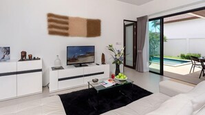 Un salon moderne doté d’un téléviseur à écran plat, d’un canapé blanc, d’une table basse en verre et d’une grande porte-fenêtre coulissante donnant sur un espace piscine.