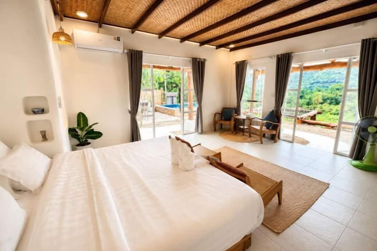A bedroom with a large bed, a wooden bench, a small table, and a view of a pool and greenery through the sliding doors.