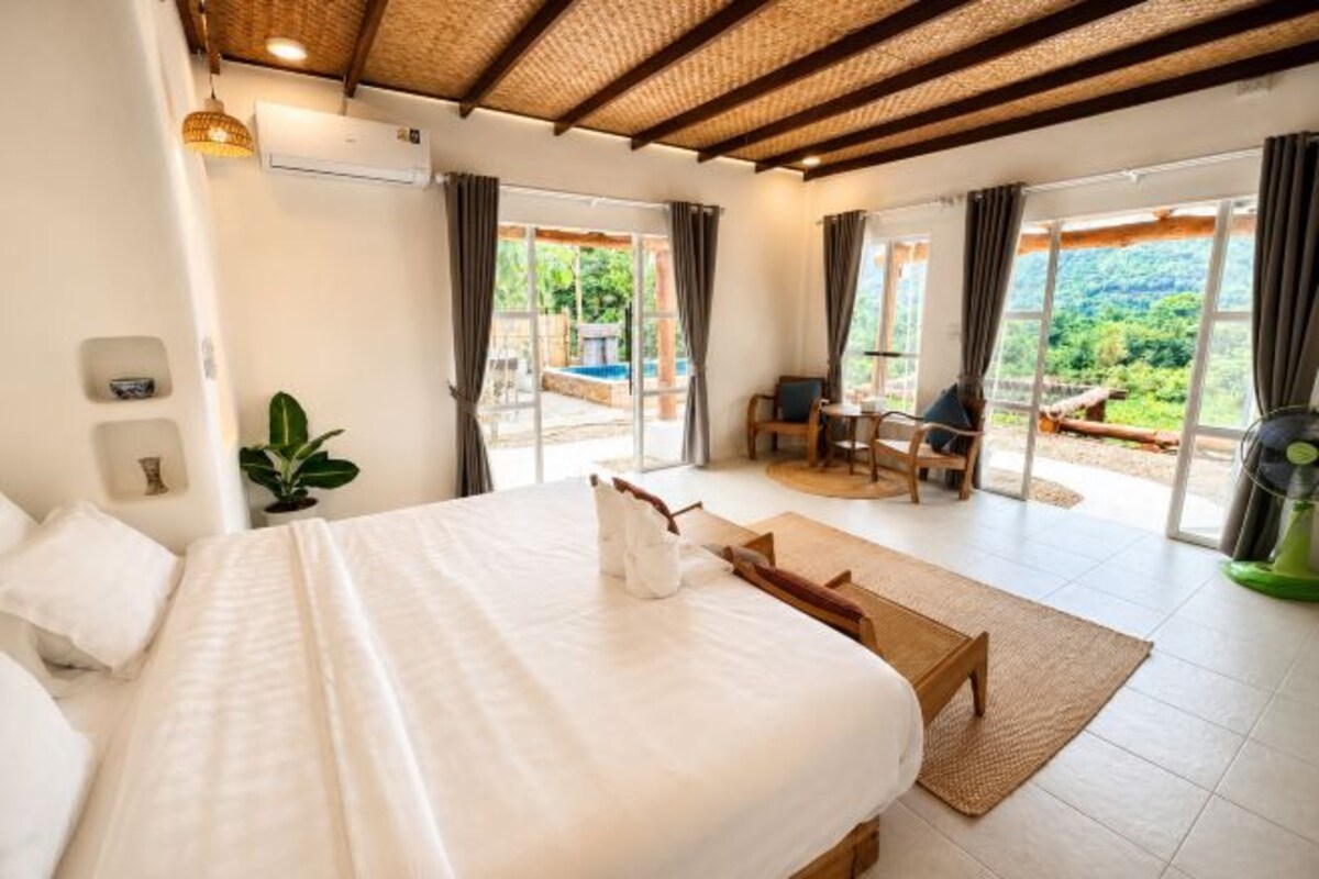 A bedroom with a large bed, a wooden bench, a small table, and a view of a pool and greenery through the sliding doors.