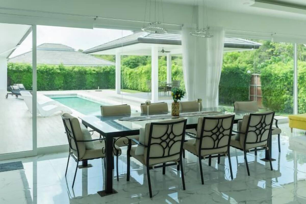 A modern dining area with a glass table and chairs, a view of a swimming pool, and a covered outdoor space.