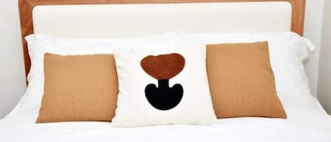 Ein Bett mit weißem Kopfteil, zwei braune Kissen und einem zentralen Kissen mit Herzmuster in Schwarz.