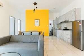 A modern living room with a sofa, a dining table, and a kitchen area with cabinets and a refrigerator.