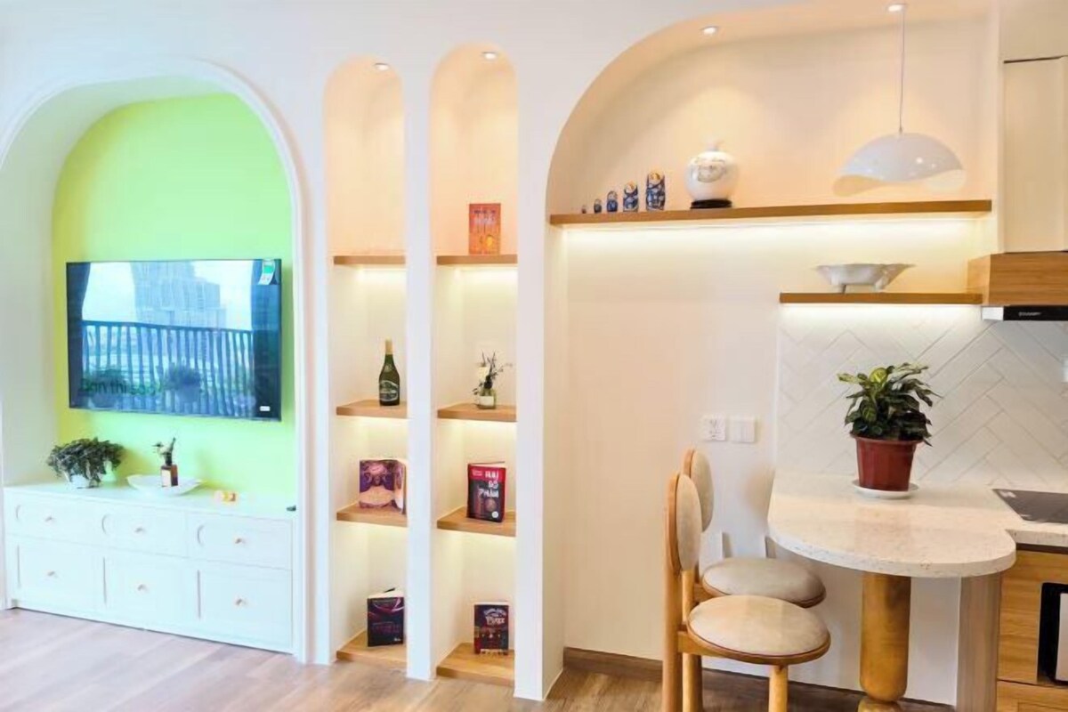 A modern kitchen with a dining area, a TV mounted on a green wall, and a potted plant on the counter.