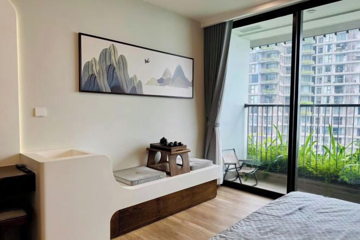 A modern hotel room with a large window, a bed, a small seating area, and a painting on the wall.