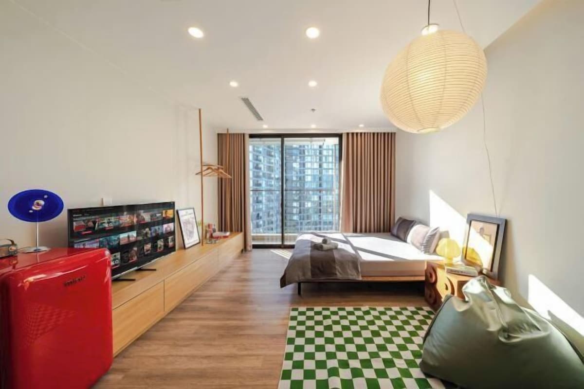 Una habitación de hotel moderna con cama, televisión, refrigerador rojo y un ventanal grande.
