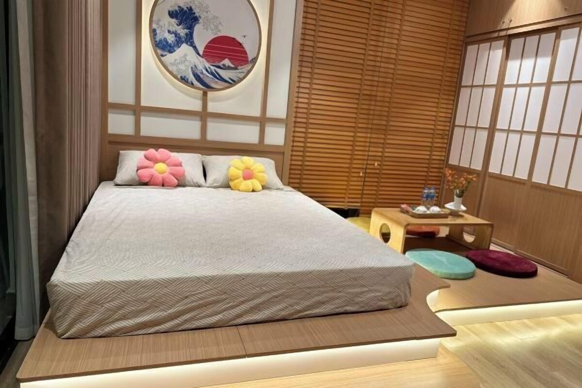 A modern bedroom with a bed, pillows, a wall-mounted artwork, a wooden side table, and a window with blinds.