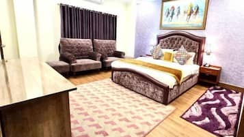 A bedroom with a bed, two armchairs, a wooden desk, a patterned rug, and a painting on the wall.
