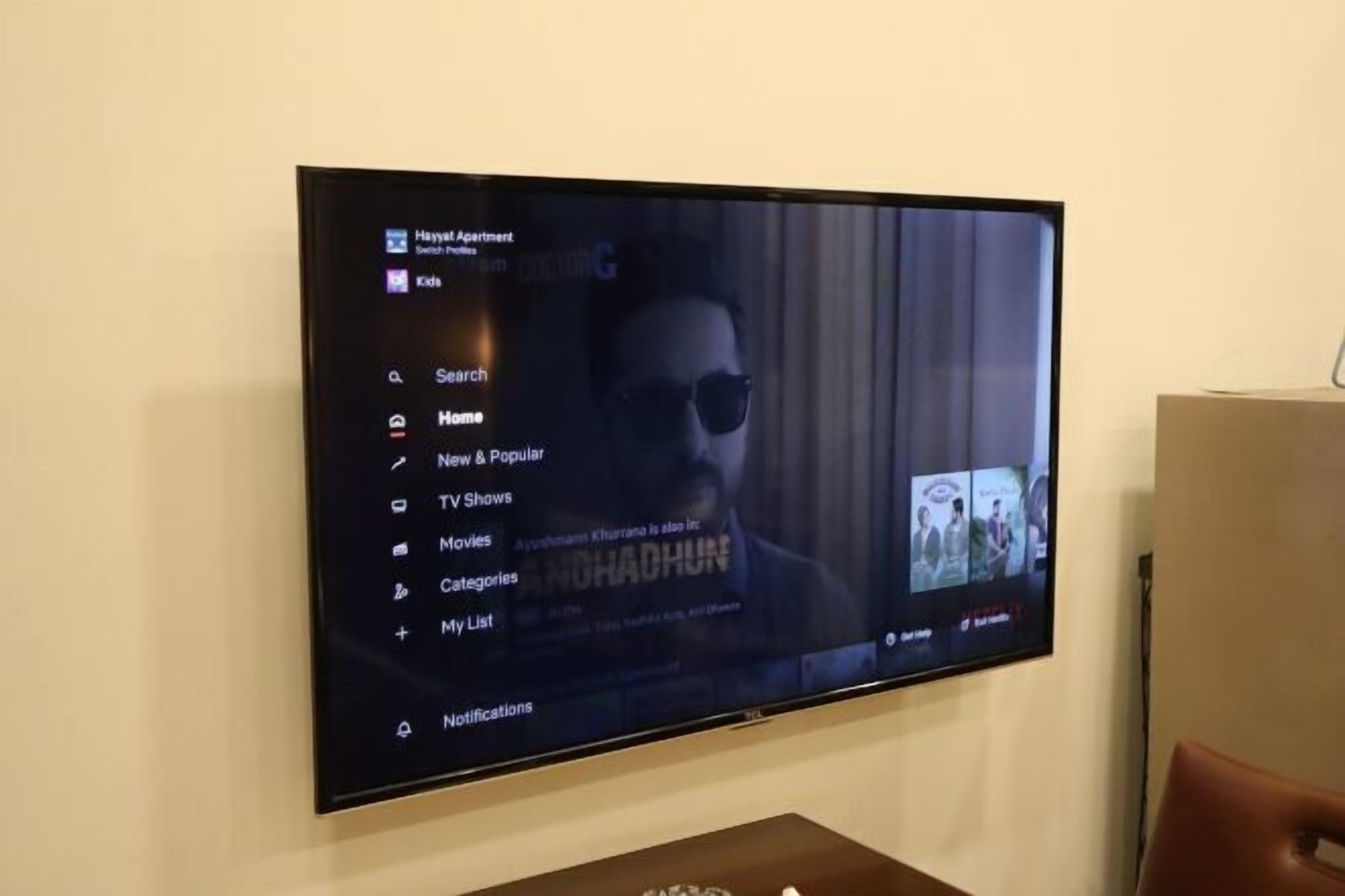 Un televisor de pantalla plana montado en una pared mostrando una interfaz de programa con opciones como “Buscar”, “Inicio”, “Novedades y populares”, “Series de televisión”, “Películas” y “Categorías”.