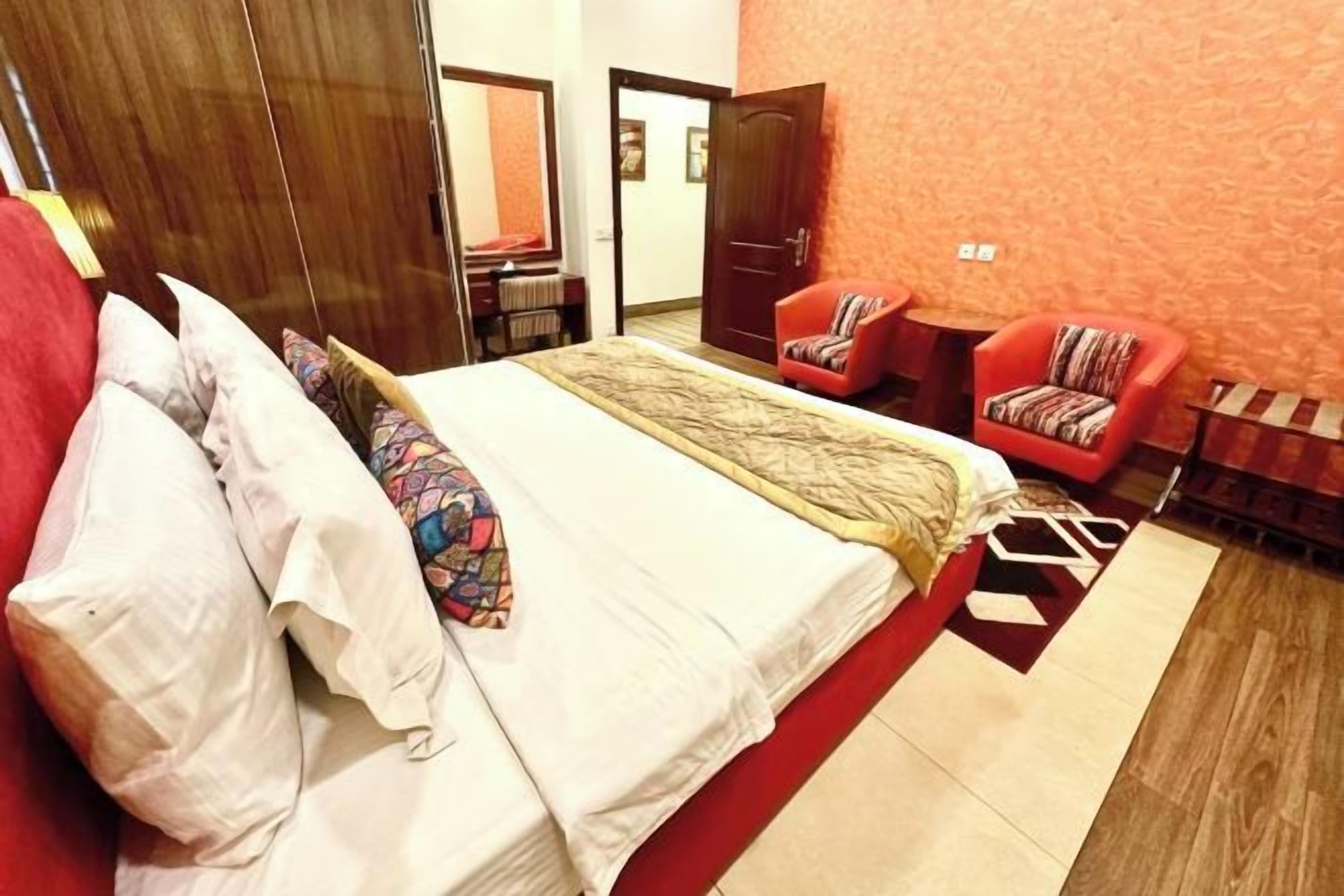 Habitación de hotel con una cama, dos sillas rojas, un armario de madera y una pared naranja con textura.