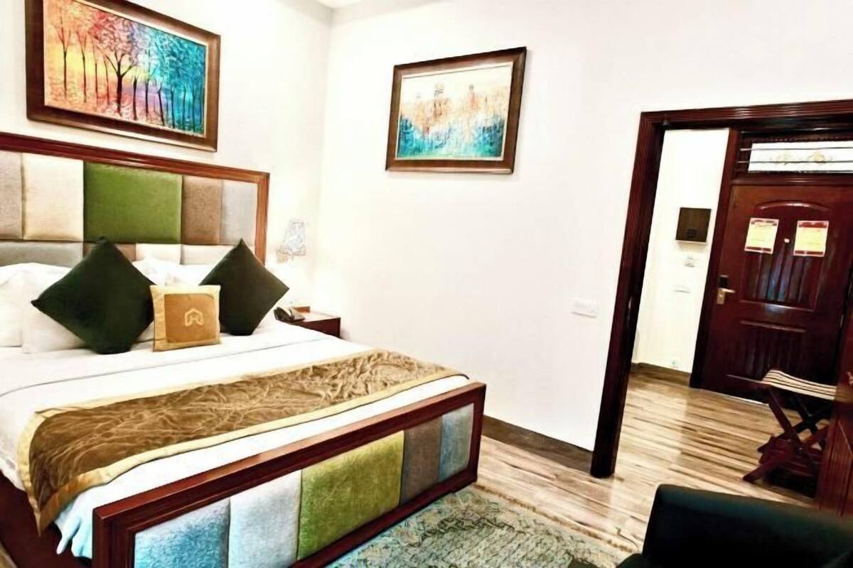 A hotel room with a large bed, a headboard with a colorful panel, a wooden door, and a small table with a lamp.