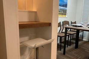 A room with a wooden shelf, a cushioned bench, a dining table with chairs, and a window with blinds. - Four Seasons Yado 1 (Tokyo)