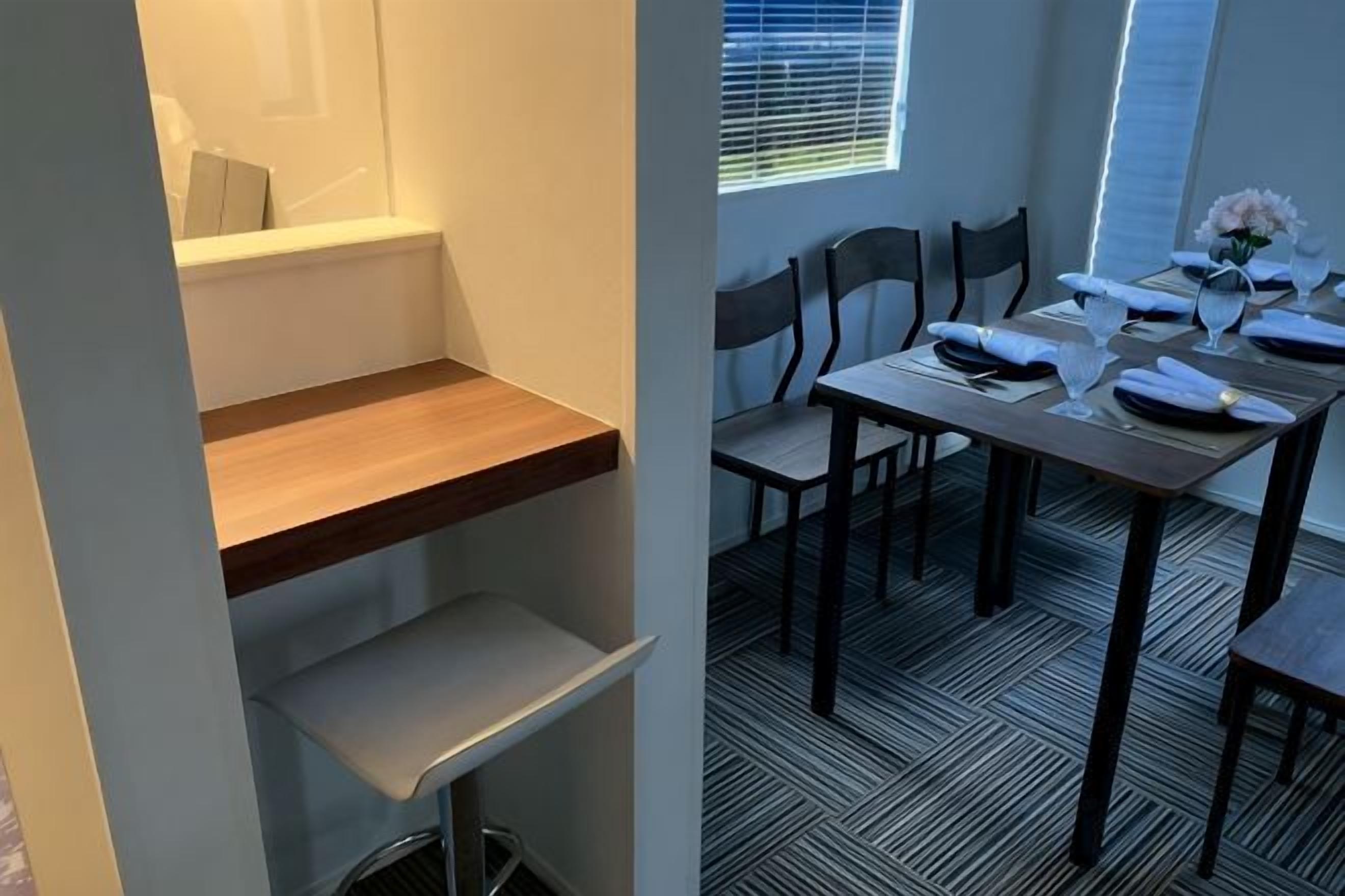 Een eetruimte met een tafel voor twee personen, stoelen, een raam met jaloezieën en een vloer met een patroon.